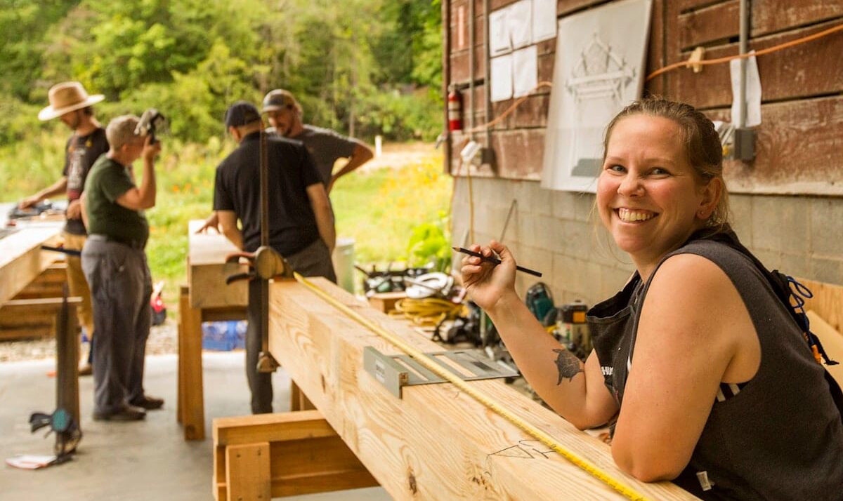 A woman smiles up from practicing measuring and cutting mortise and tenon joinery in a Timber Framing Workshop at Wild Abundance