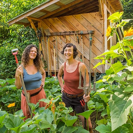 Organic vegetable gardening instructors Natalie Bogwalker and Chloe Leiberman smile holding their favorite gardening tools in the midst of a lush thriving garden at Wild Abundance