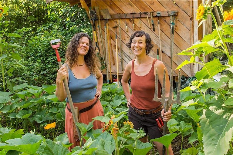 Organic vegetable gardening instructors Natalie Bogwalker and Chloe Leiberman smile holding their favorite gardening tools in the midst of a lush thriving garden at Wild Abundance