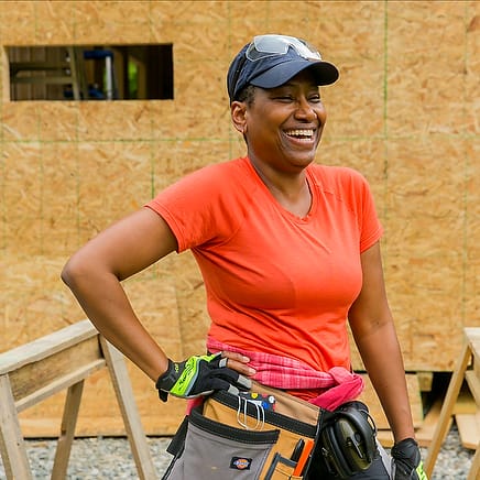 A woman smiles in front of a DIY Tiny House she's building with help from the Online Tiny House Academy, a comprehensive online program teaching each step of the building process for custom tiny homes.