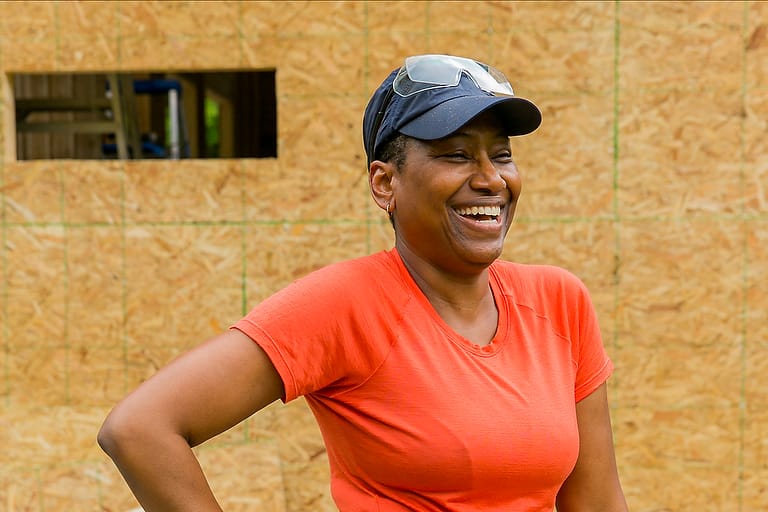 A woman smiles in front of a DIY Tiny House she's building with help from the Online Tiny House Academy, a comprehensive online program teaching each step of the building process for custom tiny homes.