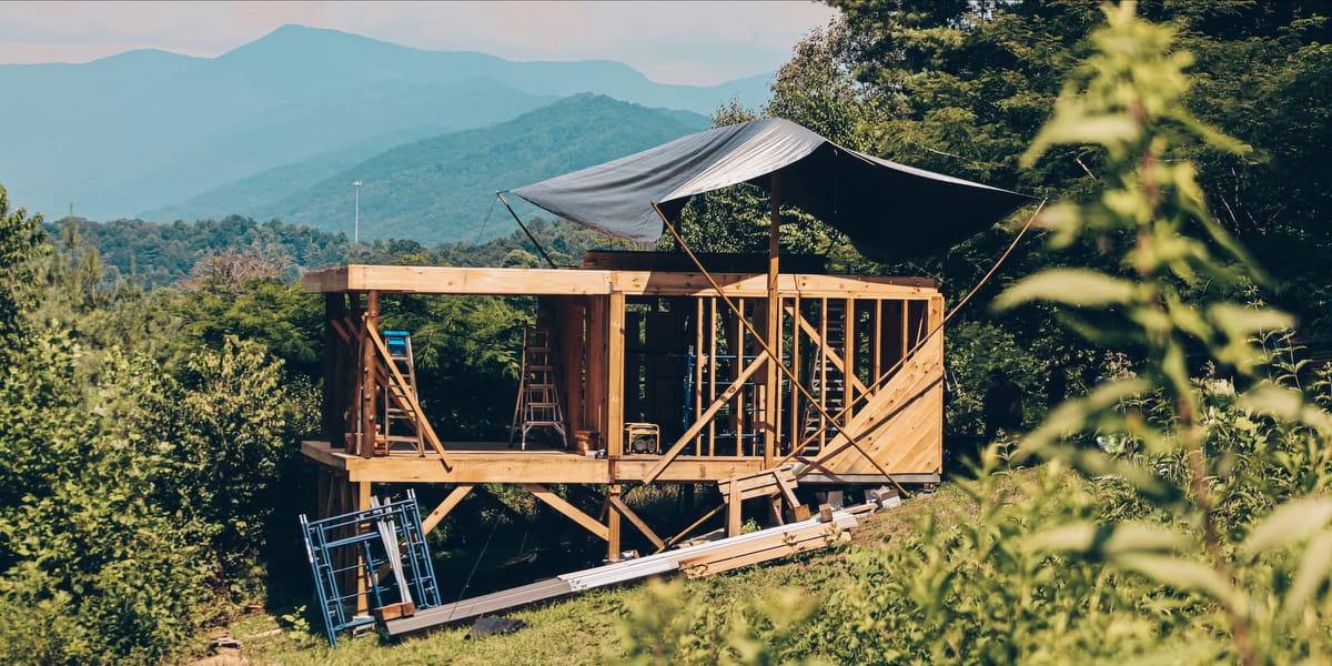 A custom DIY Tiny House being built on a hillside with a beautiful mountain view  is seen mid-construction with a tarp covering the exposed lumber 