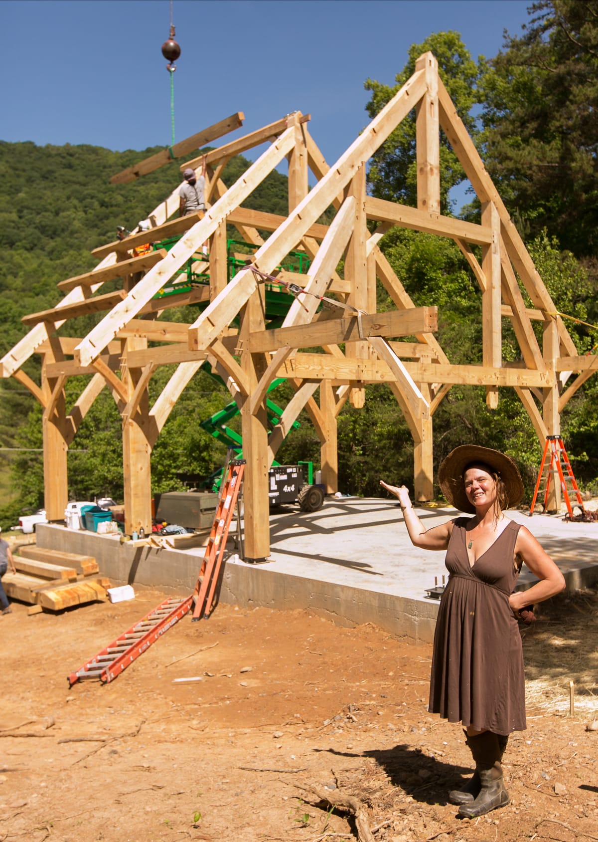 Natalie Bogwalker, founder and director of Wild Abundance, smiles and gestures to the giant timber frame teaching pavilion mid-construction behind her as it is being erected by a Timber Framing Workshop hosted at the Wild Abundance carpentry campus