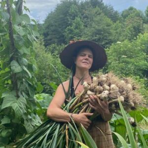 Woman stands in garden and holds large garlic harvest in arms