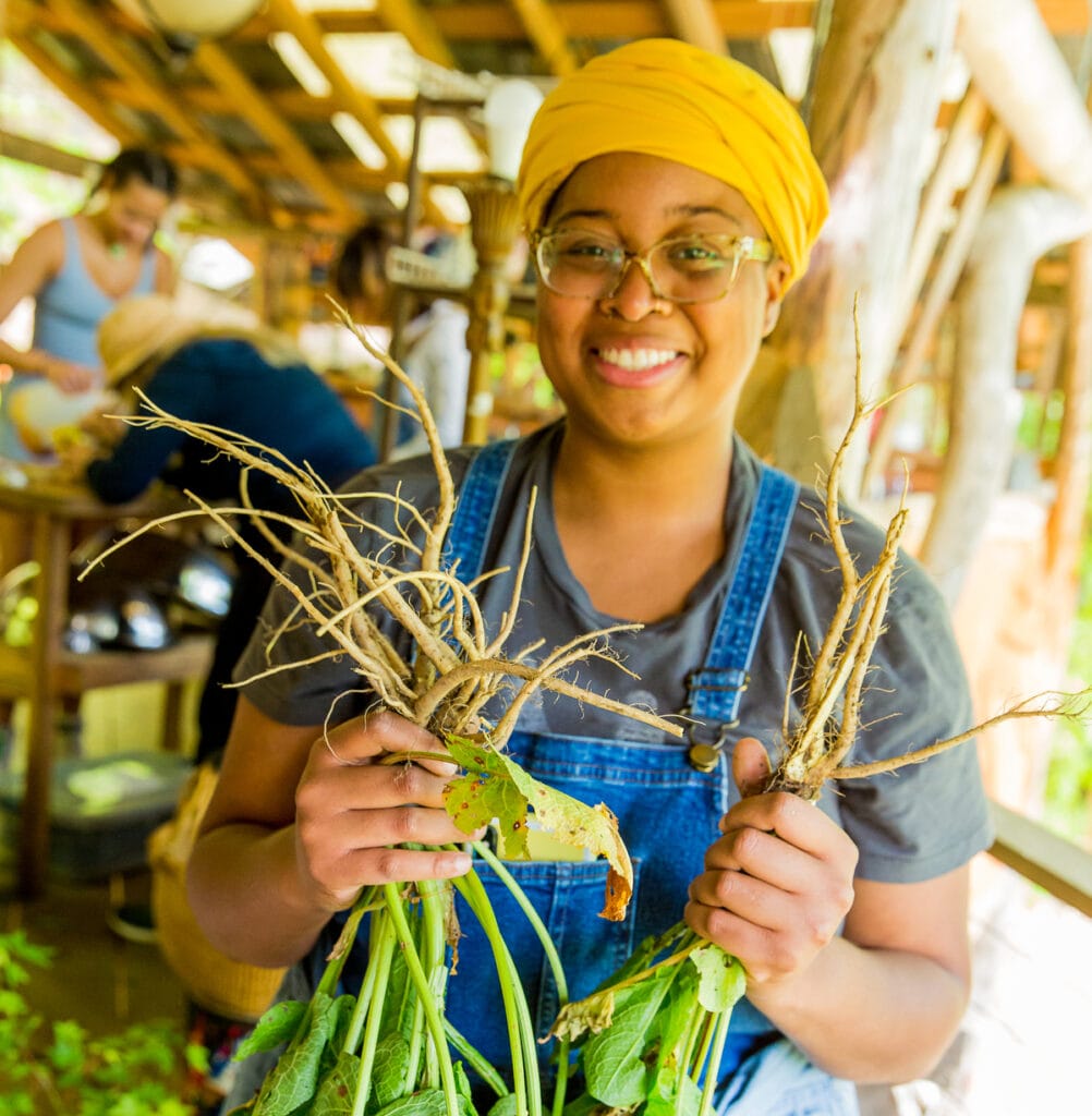 A young black woman smiles showing the roots of medicinal herbs foraged for making an herbal medicinal preparation in the Wildcrafted Apothecary 4 day workshop with Rebecca Beyer at Wild Abundance