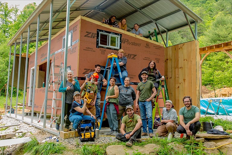 A class photo of smiling students in front of the Tiny House shell they've built during a 10 day carpentry tiny home construction workshop at Wild Abundance