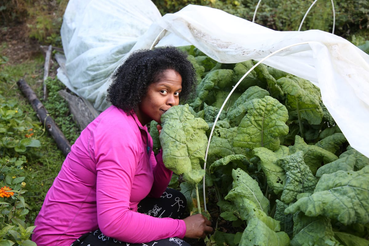 Black woman apprentice smiles up with huge kale plants in a garden under frost cloth row cover