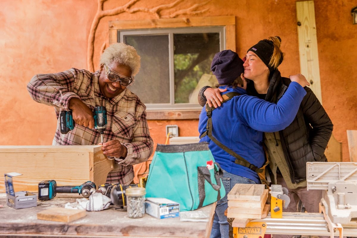 2 female students hug goodbye while another smiles as she finishes building her final project on the last day of a Women's basic carpentry workshop at Wild Abundance