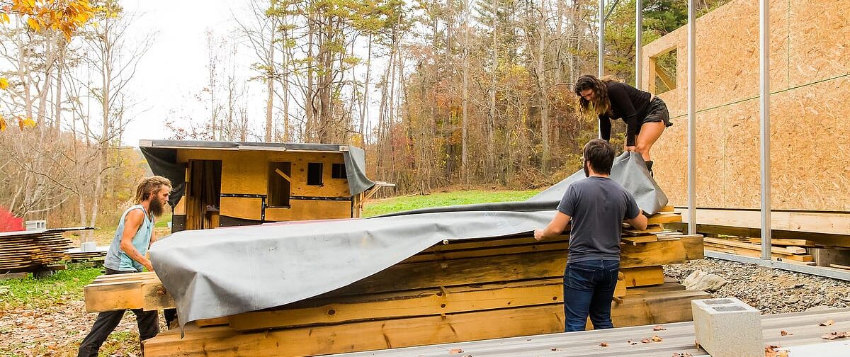 Instructor Natalie covers a stack of lumber with a tarp to prepare for a storm at the carpentry campus of Wild Abundance with 2 young men 