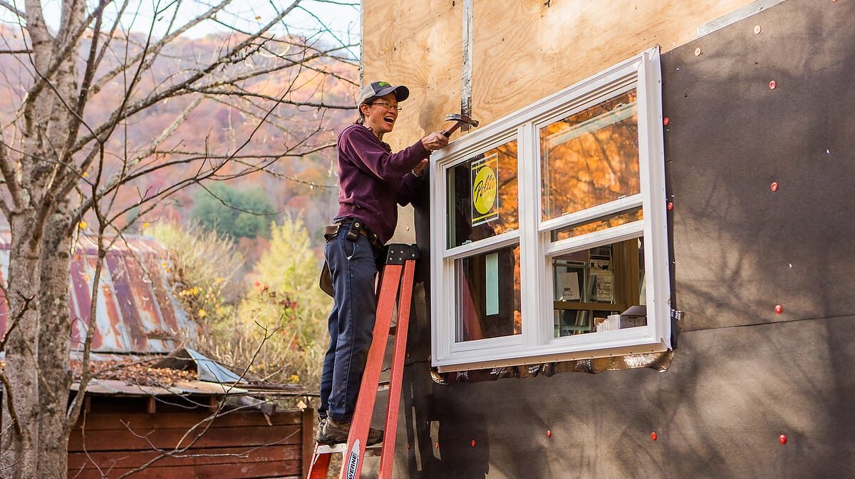 A woman smiles as she is putting the finishing touches on a window she installed during a Tiny House building workshop at Wild Abundance